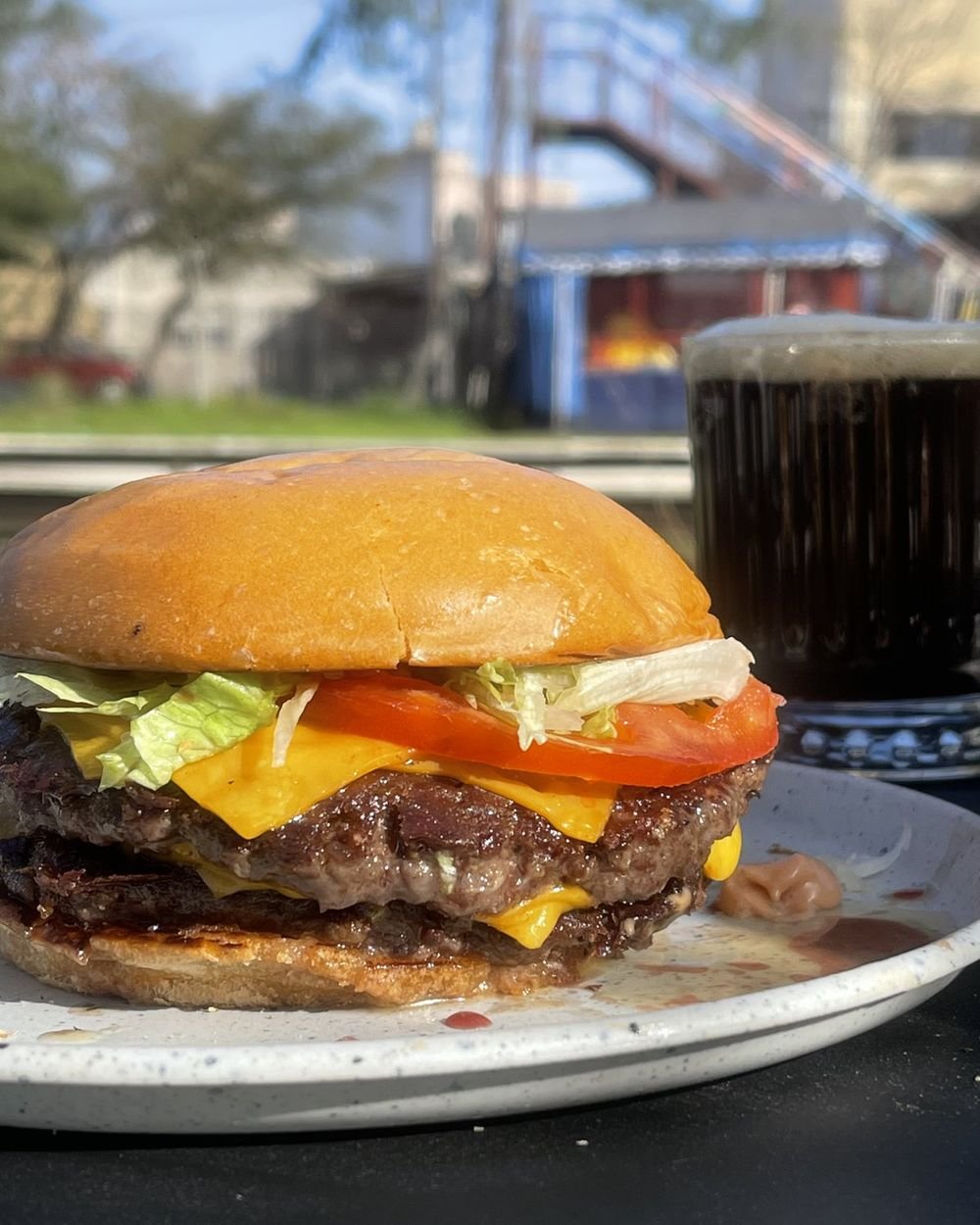 Hamburguesa doble con cheddar, tomate y lechuga en pan de papa, con una cerveza negra detrás