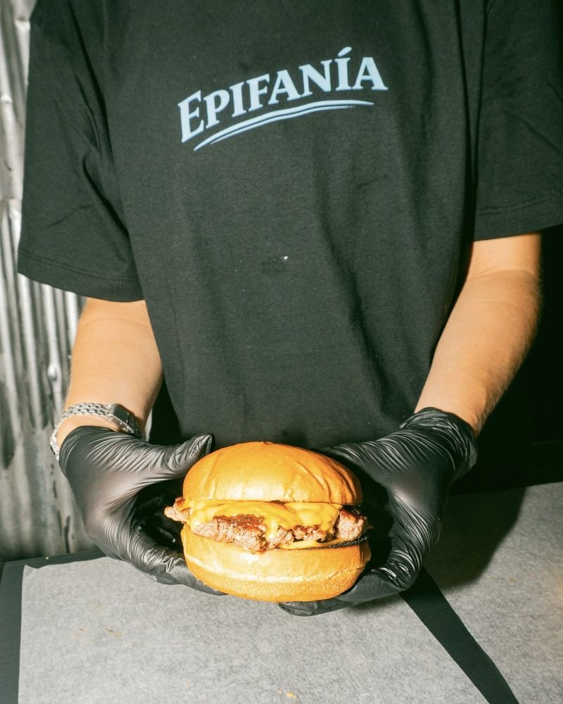 Hamburguesa sostenida con guantes negros frente a la remera con el logo de Epifanía