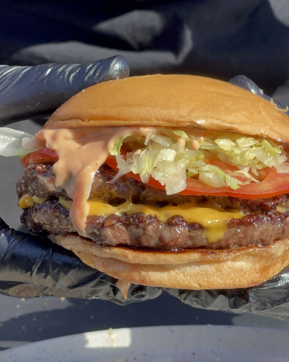 Hamburguesa doble con cheddar, tomate, lechuga y salsa chorreando, sostenida con guantes negros