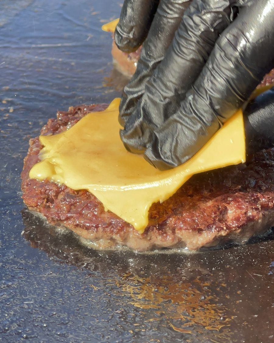 Mano con guante negro apoyando una feta de cheddar sobre la carne en la plancha